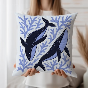 Puede incluir: Cojín cuadrado blanco con diseño marino. Dos ballenas azul oscuro con vientres azul claro nadan entre corales azul claro. Un elemento decorativo para una habitación de temática náutica u oceánica.