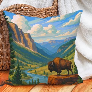May include: Square throw pillow featuring a scenic landscape with a bison. The pillow displays a vibrant painting of a mountain valley, river, and blue sky with clouds. The bison stands on a rock formation. The pillow is likely made of soft fabric.