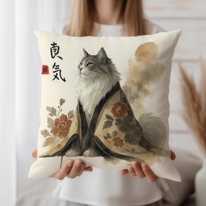 Puede incluir: Un cojín decorativo con una ilustración en acuarela de un gato con un kimono. El gato es gris y blanco con ojos verdes. El kimono es de color beige con motivos florales rojos. Hay caracteres japoneses impresos en el cojín.