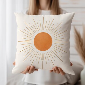 Puede incluir: Cojín cuadrado color crema con un diseño de sol. El sol es un círculo naranja con rayos dorados. El cojín es de tela suave, adecuado para una sala de estar o dormitorio. Las medidas son en centímetros.