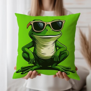 Könnte beinhalten: Ein grünes quadratisches Kissen mit einer Cartoon-Frosch-Illustration, der eine Sonnenbrille trägt. Der Frosch ist grün mit einem hellgrünen Bauch und vor einem hellgrünen Hintergrund. Die Sonnenbrille ist hellbraun.