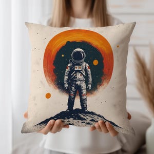 Op de afbeelding: Een vierkant decoratief kussen met een afbeelding van een astronaut die op een rotsachtig oppervlak staat, tegen een oranje en donkerblauwe hemelachtergrond. De astronaut draagt een wit ruimtepak. Het kussen heeft een vintage, ruimte-thema ontwerp.