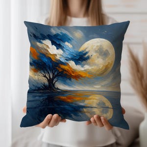 Puede incluir: Un cojín cuadrado decorativo con un diseño de paisaje artístico. La obra de arte representa un árbol con follaje azul, naranja y blanco, una gran luna y una masa de agua reflectante. El cojín tiene un fondo azul oscuro.