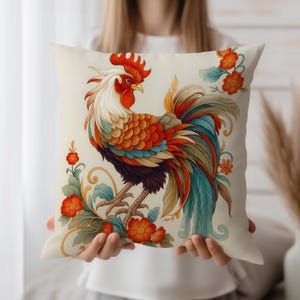 Könnte beinhalten: Quadratisches Zierkissen mit einer farbenfrohen Hahn-Illustration. Der Hahn hat rote, orange, blaue und weiße Federn mit floralen Akzenten in Orange und Grün. Das Kissen ist cremefarben.