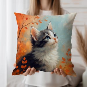 Può includere: Un cuscino decorativo con un dipinto ad acquerello di un gattino con gli occhi blu che guarda in alto. Il cuscino ha uno sfondo con tonalità arancioni e blu con fogliame autunnale.