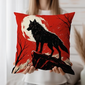May include: Red decorative pillow featuring a black wolf silhouette against a red background with a full moon. The wolf is standing on a rock, with bare tree branches on either side. A decorative item for a living room or bedroom.