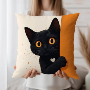 Könnte beinhalten: Quadratisches Zierkissen mit einer schwarzen Katze mit leuchtend orangefarbenen Augen und einem herzförmigen weißen Abzeichen. Die Katze lugt hinter einer orangefarbenen Tafel hervor. Das Kissen hat einen cremefarbenen Hintergrund mit orangen Sprenkeln.