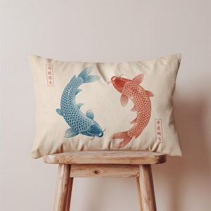 Può includere: Cuscino rettangolare beige con un disegno di due carpe koi, una blu e una rossa, che si fronteggiano. Caratteri cinesi rossi sono presenti sui lati. Il cuscino è appoggiato su uno sgabello di legno.
