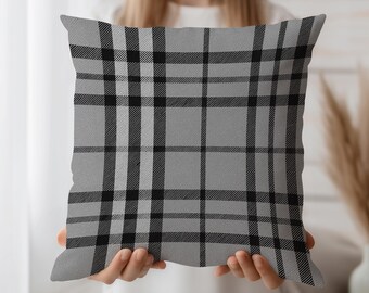 May include: A square pillow with a gray plaid pattern. The pillow features a repeating design of black and light gray lines, creating a classic checkered look. The pillow is held up by a person.