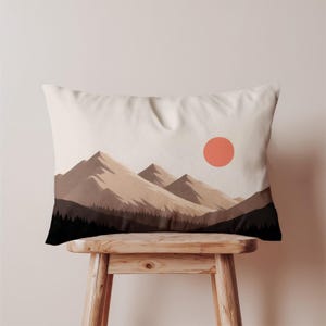 May include: Rectangular pillow with a mountain landscape design. The pillow features a stylized illustration of brown mountains, a forest, and a peach-colored sun against a cream-colored background. The pillow is resting on a wooden stool.