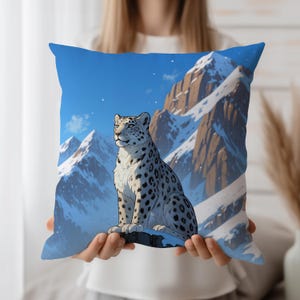 May include: A square throw pillow featuring a snow leopard sitting on a rock with a mountain range backdrop. The pillow has a blue sky and snow-covered mountains. The snow leopard has black spots and is looking to the right.