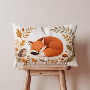May include: A rectangular pillow featuring a sleeping orange fox with a white-tipped tail. A small hedgehog and autumn leaves surround the fox. The pillow is on a wooden stool. The design has a whimsical, woodland theme.