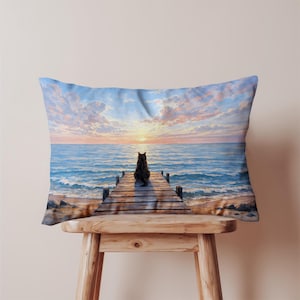 May include: Rectangular throw pillow featuring a serene beach scene. A black cat sits on a wooden pier, gazing at a sunset over the ocean. The sky is filled with pink and blue clouds. The pillow is placed on a wooden stool.