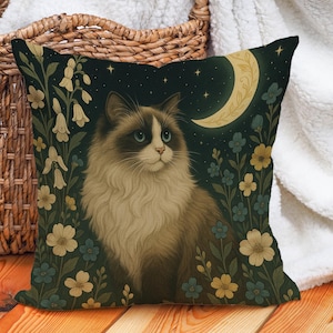 May include: Decorative square pillow featuring a cat with blue eyes, surrounded by flowers and a crescent moon against a dark background. The cat has brown and white fur. The pillow is placed near a wicker basket and a white blanket.