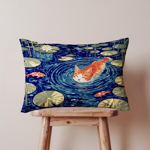 May include: Decorative rectangular pillow featuring an orange tabby cat swimming in a blue pond with lily pads and koi fish. The cat is in the center of the image, surrounded by concentric circles in the water. The pillow is on a wooden stool.