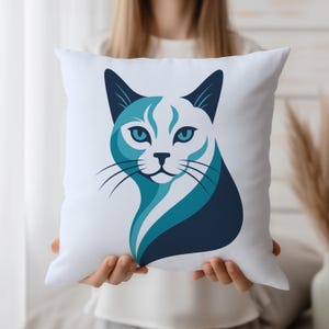 May include: White square pillow featuring a stylized cat face in shades of blue and navy. The cat's face is a graphic design with a modern aesthetic. The pillow is held up by a person.