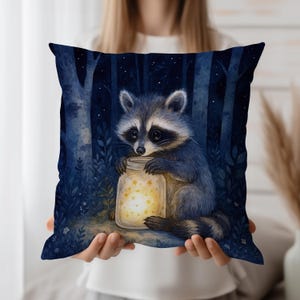 Puede incluir: Un cojín cuadrado decorativo con una ilustración de acuarela de un mapache sosteniendo un tarro lleno de estrellas brillantes. El mapache está situado sobre un fondo de bosque azul oscuro con árboles y un cielo estrellado. El cojín tiene un borde azul oscuro.