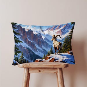 Può includere: Cuscino rettangolare con un paesaggio montano e un bighorn. Il cuscino presenta una catena montuosa innevata, alberi sempreverdi e un cielo blu. La pecora sta su una roccia coperta di neve, con le sue corna ricurve visibili. Il cuscino è posto su uno sgabello di legno.