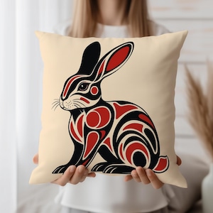 May include: A square decorative pillow with a cream-colored background. The pillow features a stylized rabbit design in black and red. The rabbit has long ears and is sitting upright, with intricate patterns on its body.