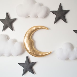 Puede incluir: Una luna creciente dorada y cinco estrellas grises de fieltro están dispuestas alrededor de dos nubes blancas de fieltro sobre un fondo blanco.