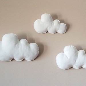 May include: Three white felt cloud shapes are attached to a light brown wall. The clouds are different sizes and are arranged in a staggered pattern.