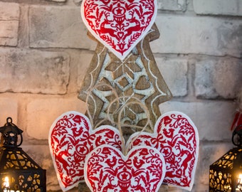 Puede incluir: Un árbol de Navidad de madera con un diseño decorativo calado sostiene varios adornos en forma de corazón. Los adornos presentan un patrón rojo y blanco con motivos de ciervos y florales. Dos faroles negros con velas encendidas flanquean el árbol.