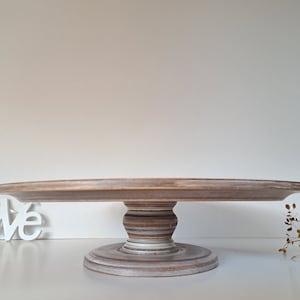 Könnte beinhalten: Ein rustikaler Kuchenständer aus Holz mit einer runden, leicht erhöhten Oberseite und einem dekorativen, gedrehten Sockel. Der Ständer ist in Braun- und Weißtönen lackiert und hat ein Used-Finish. Der Durchmesser beträgt ca. 30 cm.