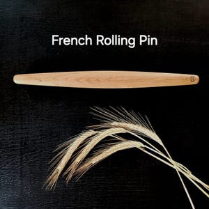 Può includere: Un mattarello francese in legno chiaro di forma arrotondata. Il testo "French Rolling Pin" è sopra il mattarello. Un fascio di spighe di grano è sotto.