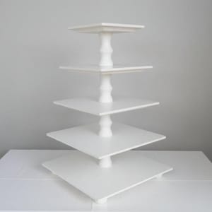 Può includere: Un'alzata quadrata bianca a cinque livelli per dolci, caratterizzata da una colonna di supporto centrale verticale con un design a gradini. Ogni livello è costituito da una piattaforma quadrata piatta che diminuisce di dimensioni dalla base alla cima.
