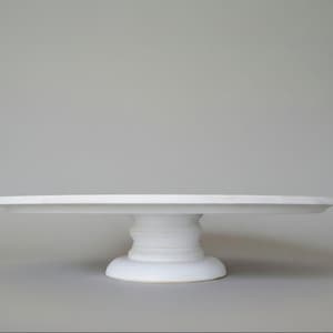 May include: A white ceramic cake stand with a wide, round top and a pedestal base. The base has a series of stacked, rounded elements. The stand is designed to display cakes and other baked goods. The background is a solid, neutral grey.