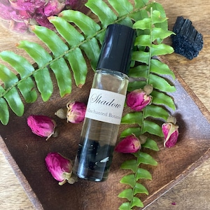 May include: A small glass bottle with a black rollerball top. The bottle is filled with a clear liquid and contains black stones. The label on the bottle reads "Shadow Enchanted Botanics".