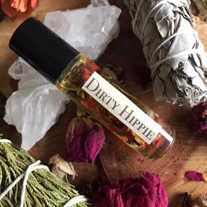 May include: A clear glass roller bottle with a black cap and a white label that reads "Dirty Hippie". The bottle is filled with a golden liquid and dried flowers. The bottle is surrounded by dried flowers, white sage, and crystals.