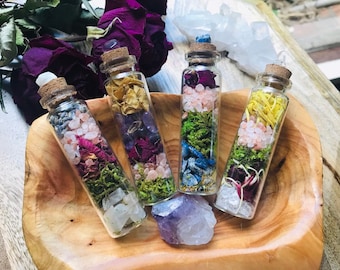 May include: Five small glass bottles filled with dried flowers, herbs, and crystals. The bottles have cork stoppers and are arranged in a wooden bowl.