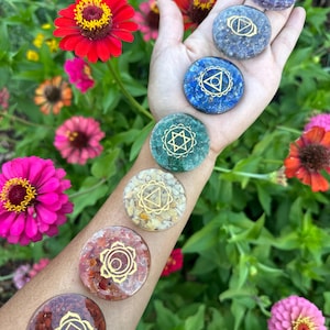May include: Seven chakra stones with gold symbols on a hand. The stones are in the colors of the chakras: red, orange, yellow, green, blue, indigo, and violet.