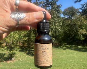 Op de afbeelding: Een bruine glazen fles met een zwarte druppelaar, met het opschrift "Yarrow" en "Enchanted Botanicals". De fles wordt vastgehouden tegen een achtergrond van groen gras en bomen onder een blauwe lucht. Het etiket heeft een vintage esthetiek.
