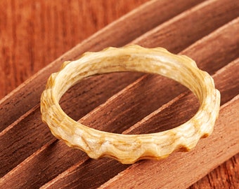 May include: A light brown wooden ring with a textured, organic design. The ring is lying on a wooden surface.