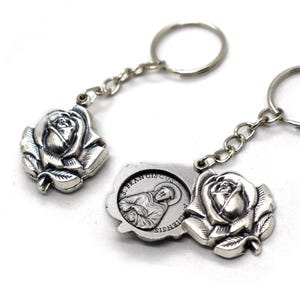 May include: Two silver keychains with rose-shaped pendants. One keychain features a small, rectangular pendant with an image of a saint and the text "S. FRANCISCUS SISNENSIS". The keychains have silver chain links and key rings.