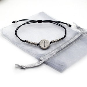 Puede incluir: Un brazalete de cuerda negra con cuentas plateadas y un dije plateado con una medalla de San Benito. El brazalete se muestra sobre un fondo blanco con una bolsa de cordón plateada.