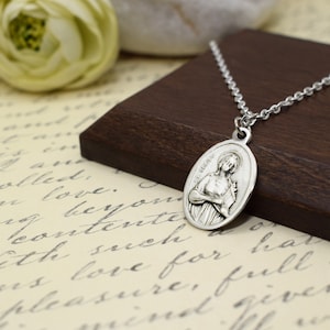 Può includere: Collana con ciondolo ovale in argento con una raffigurazione di Santa Cecilia che tiene uno strumento musicale. Il ciondolo è su una catena d'argento e poggia su una superficie di legno.