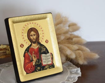 May include: A religious icon featuring a depiction of Jesus Christ, with a gold leaf background and a black frame. The icon shows Jesus in a red robe and black cloak, holding a book. The image is set on a white lace doily.