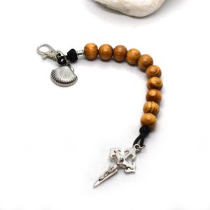 Puede incluir: Un llavero de cuentas de madera con un colgante de cruz plateada y un dije de concha. Las cuentas son de color marrón claro y están ensartadas en un cordón negro. El llavero tiene un cierre plateado.
