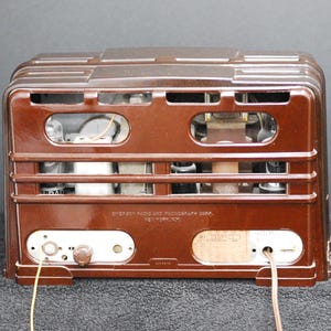 May include: A vintage brown radio with a rectangular design. The back of the radio features a series of horizontal bars, openings, and internal components. The radio has a dial, knobs, and a label with text. Wires extend from the bottom.