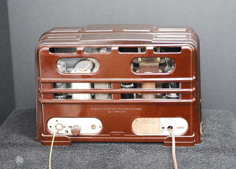 May include: A vintage brown radio with a rectangular design. The back of the radio features a series of horizontal bars, openings, and internal components. The radio has a dial, knobs, and a label with text. Wires extend from the bottom.