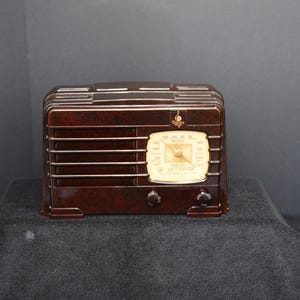 May include: A vintage brown radio with a cream-colored dial and two knobs. The radio has horizontal lines across the front and a small emblem above the dial. A classic piece of home decor.