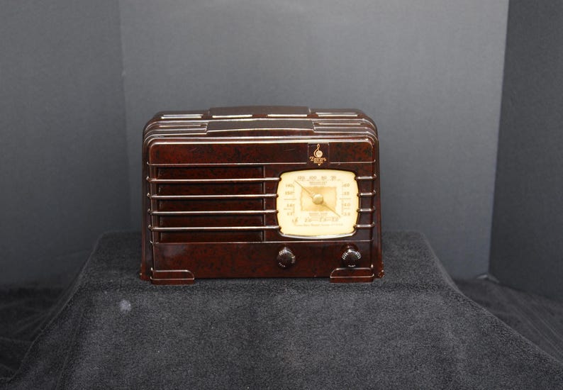 May include: A vintage brown radio with a cream-colored dial and two knobs. The radio has horizontal lines across the front and a small emblem above the dial. A classic piece of home decor.
