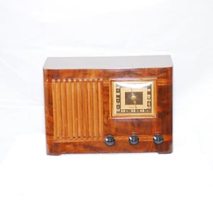 May include: A vintage wooden radio with a rectangular shape and a glossy finish. The radio has a square dial with a gold-coloured frame and black knobs. The front features vertical wooden slats.