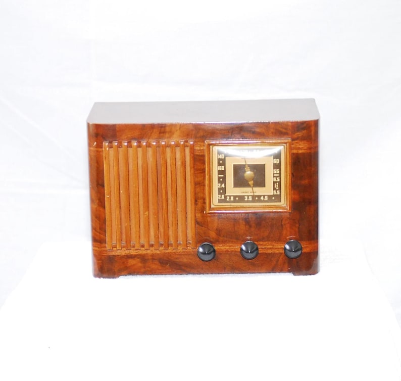 May include: A vintage wooden radio with a rectangular shape and a glossy finish. The radio has a square dial with a gold-coloured frame and black knobs. The front features vertical wooden slats.