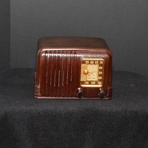 May include: A vintage brown radio with a rectangular shape. The radio has a dark wood finish with vertical lines on the front and a dial with numbers. Two black knobs are visible below the dial.