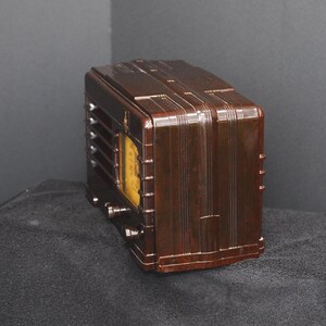 May include: A vintage brown radio with a rectangular shape and a curved top. The radio has a yellow dial and several knobs. The front of the radio has vertical lines and a speaker grill. The radio is sitting on a dark surface.