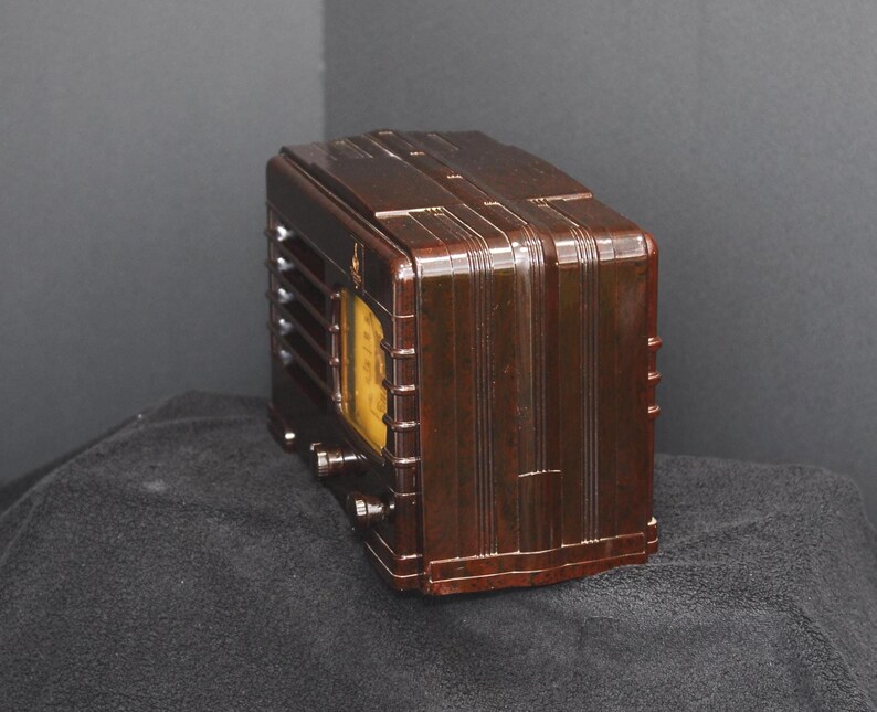 May include: A vintage brown radio with a rectangular shape and a curved top. The radio has a yellow dial and several knobs. The front of the radio has vertical lines and a speaker grill. The radio is sitting on a dark surface.
